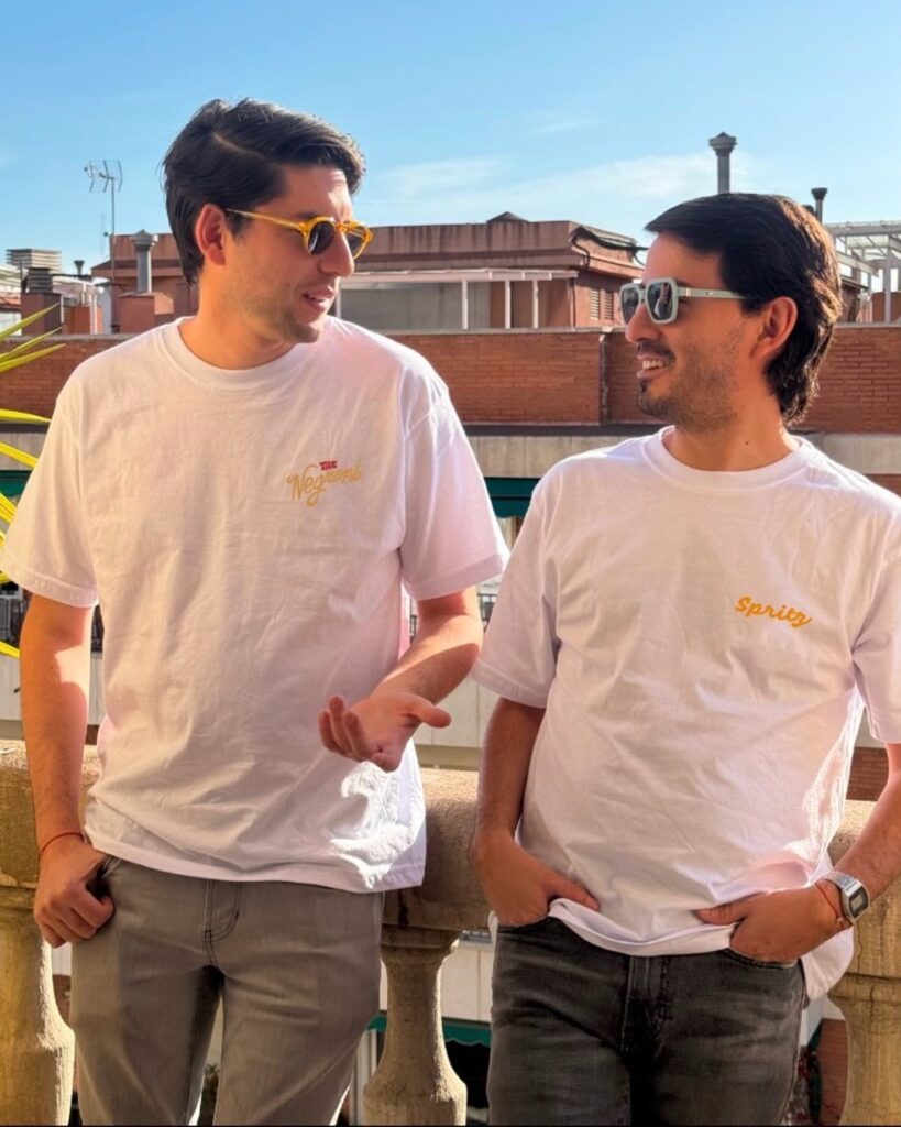 Dos amigos con las camisetas blancas The Negroni y The Aperol Spritz de agua sal en un balcón